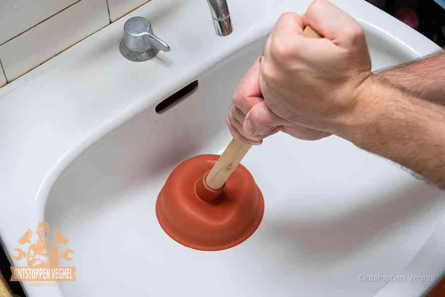 Twee handen gebruiken een rode rubberen plopper op het afvoerputje om de gootsteen te ontstoppen in Veghel