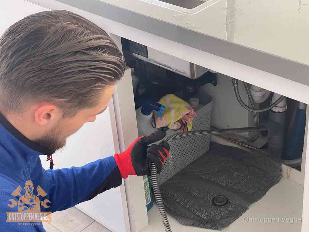 Een monteur voert een flexibele ontstoppingsveer in onder het keukenkastje, langs de sifon, om de gootsteen te ontstoppen in Veghel