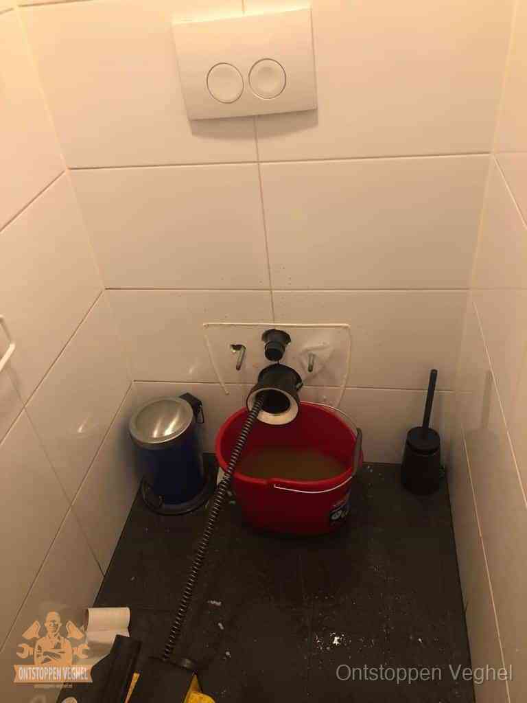 Waar zit de wc-verstopping: pot, bocht of riool in Veghel Een ontstopte wand-wc met verwijderde toiletpot waar een ontstoppingsveer in de wanduitgang verdwijnt, het achterhalen waar de verstopping zit in Veghel